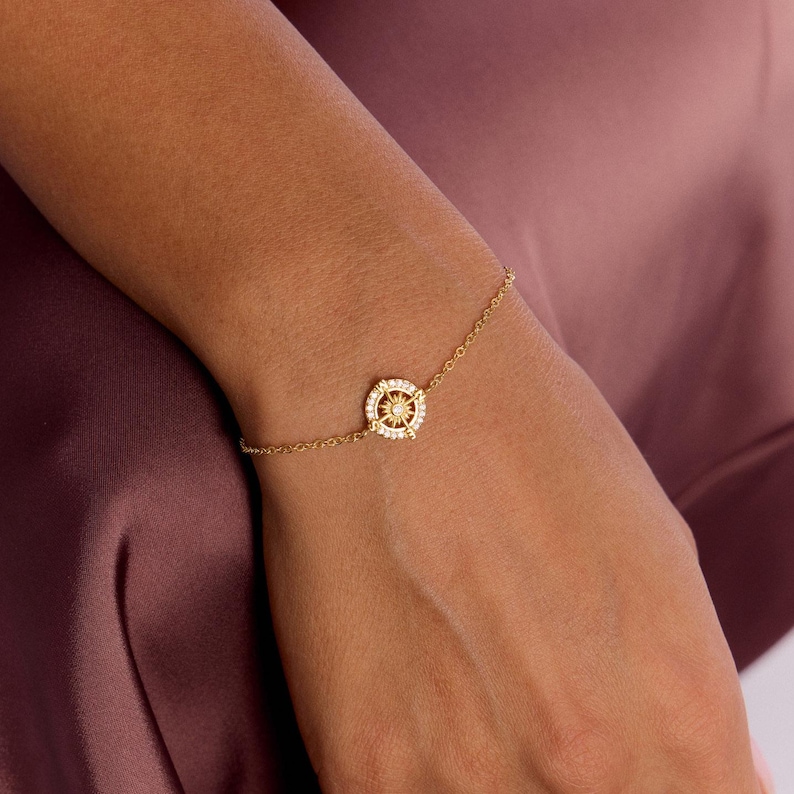 May include: A gold-colored bracelet featuring a compass rose design. The compass rose is encrusted with small, clear gemstones and is connected to a delicate chain. The bracelet is worn on a wrist.