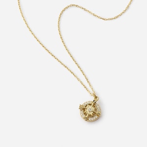 K&ouml;nnte beinhalten: Eine goldfarbene Halskette mit einem Kompass-Anh&auml;nger. Der Anh&auml;nger zeigt ein Kompassrosen-Design mit einem zentralen Stern und ist von kleinen, klaren Edelsteinen umgeben. Die Kette ist filigran und ebenfalls goldfarben.