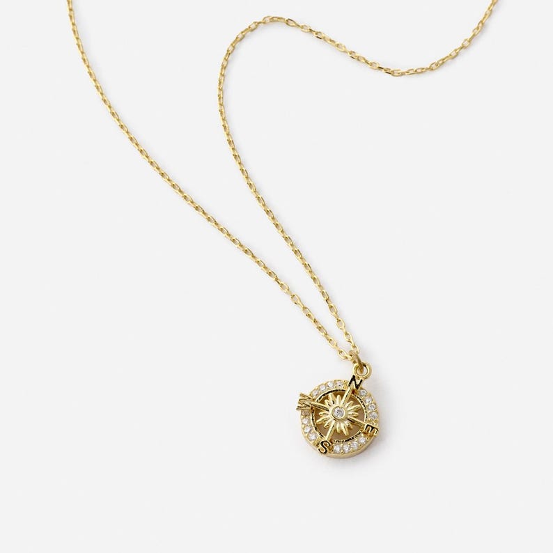 K&ouml;nnte beinhalten: Eine goldfarbene Halskette mit einem Kompass-Anh&auml;nger. Der Anh&auml;nger zeigt ein Kompassrosen-Design mit einem zentralen Stern und ist von kleinen, klaren Edelsteinen umgeben. Die Kette ist filigran und ebenfalls goldfarben.