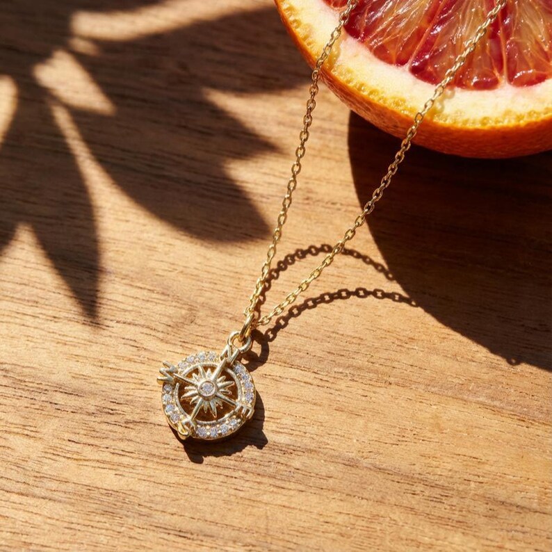 K&ouml;nnte beinhalten: Eine goldfarbene Kompass-Anh&auml;nger-Halskette mit einer filigranen Kette. Der Anh&auml;nger hat ein Kompass-Design mit einem zentralen Stern und einem Ring aus kleinen, klaren Steinen. Die Halskette liegt auf einer Holzoberfl&auml;che, mit einer Orangenscheibe im Hintergrund.