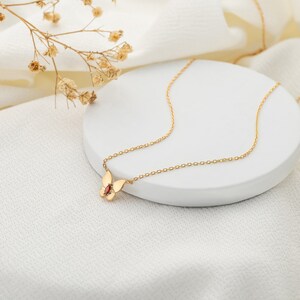 May include: A gold chain necklace with a delicate butterfly pendant featuring a red gemstone. The necklace is displayed on a white circular surface.