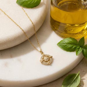 K&ouml;nnte beinhalten: Eine goldfarbene Kompass-Anh&auml;nger-Halskette mit einer zarten Kette. Das Kompass-Design hat einen zentralen Stern, der von kleinen, klaren Edelsteinen umgeben ist. Die Halskette liegt auf einer wei&szlig;en Marmoroberfl&auml;che, mit frischen Basilikumbl&auml;ttern und einer &Ouml;lflasche im Hintergrund.