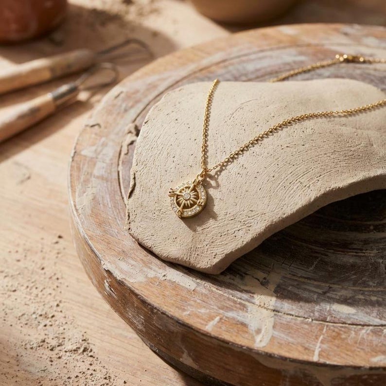 K&ouml;nnte beinhalten: Eine goldfarbene Halskette mit einem Kompass-Anh&auml;nger. Der Anh&auml;nger hat ein Sonnenstrahlen-Design und kleine, klare Steine. Die Kette liegt auf einem hellen, strukturierten Stein, der auf einer Holzoberfl&auml;che platziert ist. Der Anh&auml;nger hat einen Durchmesser von etwa 2 cm.