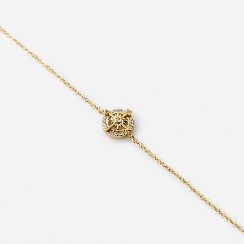 May include: A gold-colored bracelet featuring a compass design. The compass is encrusted with small, clear gemstones and has a central starburst design. The bracelet chain is delicate and also gold-colored.