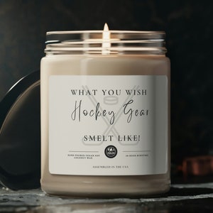 May include: A white candle with a black and white label that reads "What you wish Hockey Gear Smelt Like!" The candle is in a clear glass jar with a black lid. The candle is lit and the flame is visible.