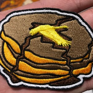 May include: A brown and yellow embroidered patch depicting a stack of pancakes with a yellow bird flying above. The patch has a white outline.