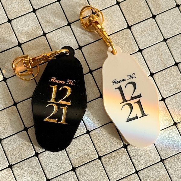 May include: Two retro hotel key fobs, one black, one cream. Both have gold-tone key rings and the text "Room No. 12 21" in gold. They are displayed on a white and grey tiled surface.