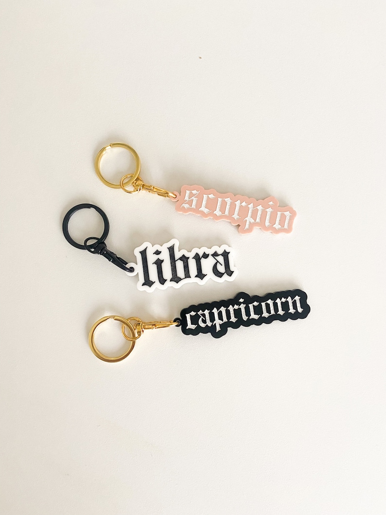 May include: Three gold keychains with different zodiac signs: Libra, Scorpio, and Capricorn. The signs are made of black and pink acrylic with white lettering.