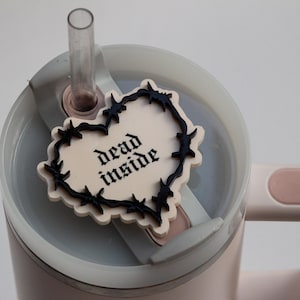 Peut inclure: Une tasse de voyage blanche avec un charme en forme de cœur noir qui dit "dead inside" dans une police gothique. Le charme est attaché au couvercle de la tasse avec un design de fil de fer barbelé noir.