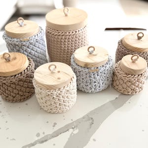 Puede incluir: Colección de tarros de almacenamiento de ganchillo hechos a mano en varios tamaños y colores neutros, incluyendo beige, gris y crema. Cada tarro presenta una tapa de madera con un pequeño asa de madera. Los tarros están dispuestos sobre una superficie blanca.