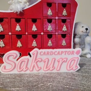 Può includere: Un cartello in legno rosa e bianco con il testo "CARDCAPTOR Sakura" in lettere rosa. Il cartello è davanti a un calendario dell'avvento in legno rosso con 24 piccoli cassetti.