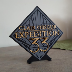 Puede incluir: Placa gris y negra en forma de diamante con las palabras "Clair Obscur Expedition 33" en letras doradas. La placa está montada sobre un soporte negro y se exhibe sobre una superficie de madera. El diseño es de estilo art déco.