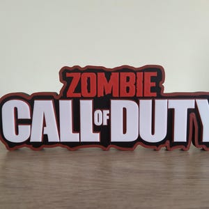 Peut inclure: Un logo "Zombie Call of Duty" rouge et blanc sur fond noir. Le logo est fait d'un matériau imprimé en 3D.