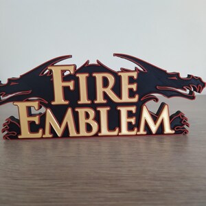 Op de afbeelding: Een 3D-geprint Fire Emblem-logo met twee zwarte draken aan weerszijden van de gouden tekst "FIRE EMBLEM". De draken hebben rode contouren.