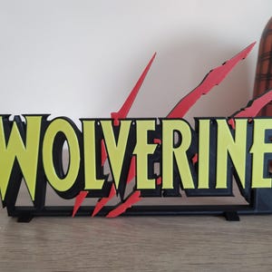 Peut inclure: Une enseigne en 3D imprimée en noir et jaune avec le mot "WOLVERINE" en lettres jaunes et des griffes rouges.