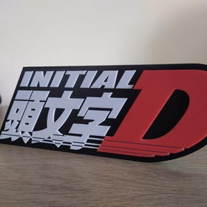 Peut inclure: Logo Initial D noir et rouge avec inscription blanche et caractères japonais. Le logo est sur fond noir et comporte un D rouge avec des rayures blanches.