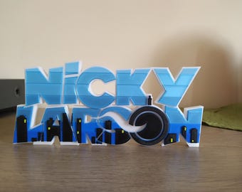 May include: A 3D printed sign with the name "Nicky Larson" in blue and white letters. The letters are layered and have a city skyline design behind them. The sign is on a wooden surface.