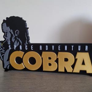 Może przedstawiać: Czarno-złoty znak z napisem "SPACE ADVENTURE COBRA" w białych i złotych literach. Znak przedstawia sylwetkę osoby palącej papierosa. Znak znajduje się na drewnianej powierzchni.