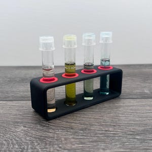 May include: A black test tube rack with four clear tubes, each containing a different colored liquid. The rack has red inserts for the tubes. The liquids are clear, yellow-green, gray, and blue-green.
