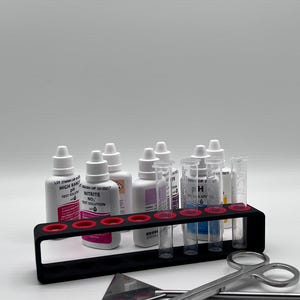 May include: A black test tube rack holds several bottles and test tubes. The bottles have white caps and labels with red and black text. A pair of silver scissors and a small metal tool are in the foreground.