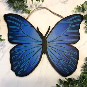 Puede incluir: Adorno de pared de mariposa de madera con un diseño azul y negro. La mariposa tiene un degradado de tonos azules en sus alas, con contornos negros y un cuerpo negro. Se adjunta un lazo de cuerda natural para colgar.
