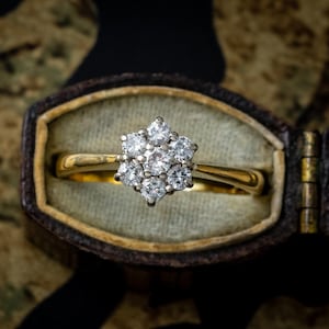 May include: A gold ring with a cluster of diamonds in a floral design. The ring is set in a vintage-style jewelry box with a velvet interior. The diamonds are clear and sparkling, and the gold band is polished.