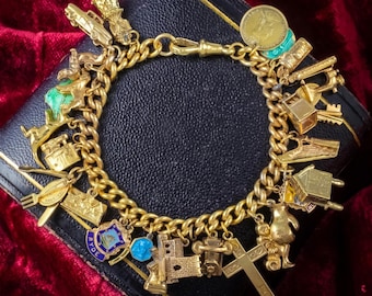May include: A gold-tone charm bracelet featuring a variety of dangling charms including a car, a coin, a cross, a house, a boxing glove, and a shield-shaped emblem with the text RYDE. The bracelet has a thick curb chain and a secure clasp.