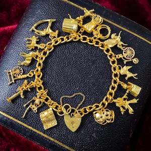 Puede incluir: Una pulsera de dijes dorada que presenta una variedad de colgantes, incluyendo un candado en forma de corazón, máscaras teatrales, un esqueleto, una rueca, un carruaje y varias figuras de animales, unidos a una banda de cadena de eslabones dorados.