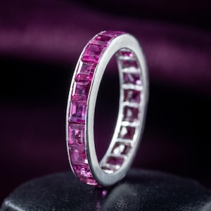 Può includere: Un anello d'argento con una fascia di pietre preziose rosa vivaci, tagliate a forma quadrata. L'anello è posto su uno sfondo scuro, che mette in risalto la brillantezza delle pietre. Un gioiello.