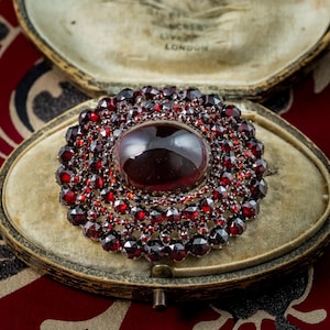 Op de afbeelding: Een vintage ovale broche met een grote, donkerrode cabochon in het midden, omringd door meerdere lagen kleinere, gefacetteerde rode edelstenen. De broche wordt gepresenteerd in een vintage, met fluweel gevoerde doos. De doos heeft de tekst "LONDON".