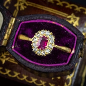 Peut inclure: Une bague en or avec une pierre précieuse rouge rectangulaire centrale entourée d'un halo de douze petites pierres rondes transparentes. La bague est placée dans un écrin marron foncé avec un intérieur en velours violet contrastant.