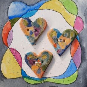 May include: Three ceramic heart-shaped buttons with a colorful abstract design. The buttons are painted with shades of blue, green, yellow, orange, and purple. Each button has a hole in the center for sewing.