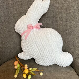 K&ouml;nnte beinhalten: Ein wei&szlig;er Pl&uuml;schhase mit einer rosa Schleife. Der Hase sitzt auf einer braunen Stoffoberfl&auml;che.