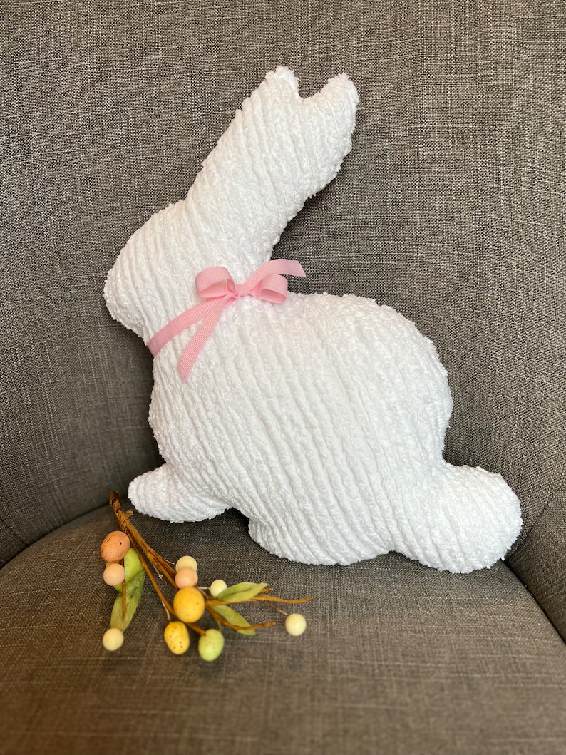 K&ouml;nnte beinhalten: Ein wei&szlig;er Pl&uuml;schhase mit einer rosa Schleife. Der Hase sitzt auf einer braunen Stoffoberfl&auml;che.