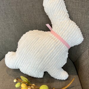 K&ouml;nnte beinhalten: Ein wei&szlig;er Pl&uuml;schhase mit einem rosa Band um den Hals. Der Hase sitzt auf einer grauen Stoffoberfl&auml;che.