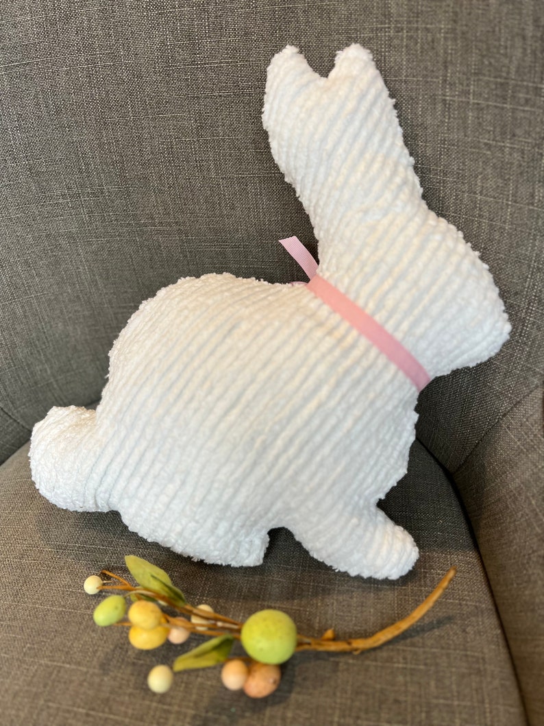 K&ouml;nnte beinhalten: Ein wei&szlig;er Pl&uuml;schhase mit einem rosa Band um den Hals. Der Hase sitzt auf einer grauen Stoffoberfl&auml;che.