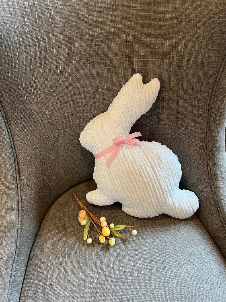 May include: A white plush bunny with a pink ribbon on its neck. The bunny is sitting on a gray chair with a sprig of green leaves and small, colorful eggs.
