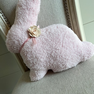 May include: A pink fluffy bunny pillow with a white flower accent. The pillow is sitting on a light gray chair.