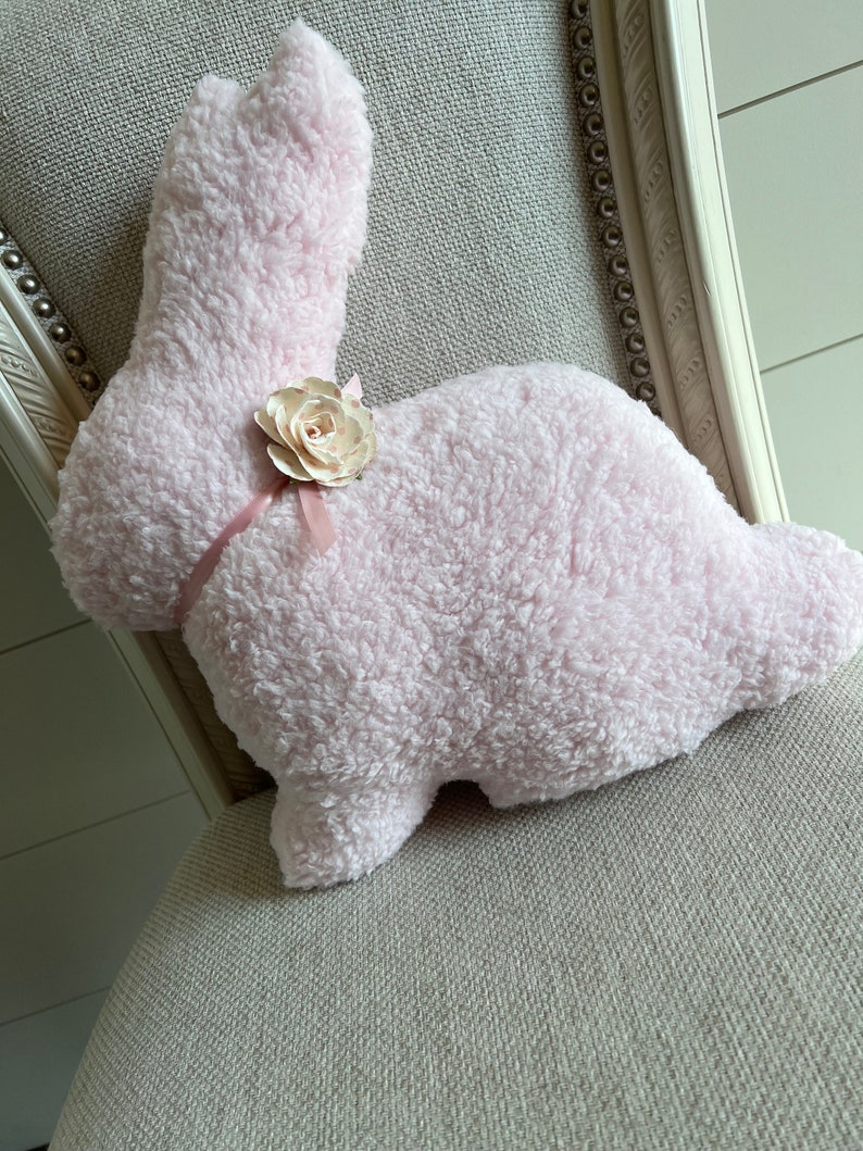 May include: A pink fluffy bunny pillow with a white flower accent. The pillow is sitting on a light gray chair.