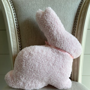 May include: A pink plush bunny rabbit pillow with a pink satin ribbon bow. The pillow is sitting on a light gray chair.
