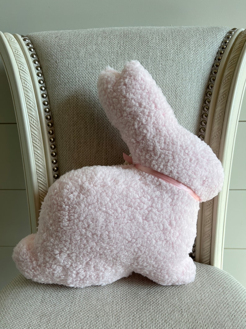 May include: A pink plush bunny rabbit pillow with a pink satin ribbon bow. The pillow is sitting on a light gray chair.