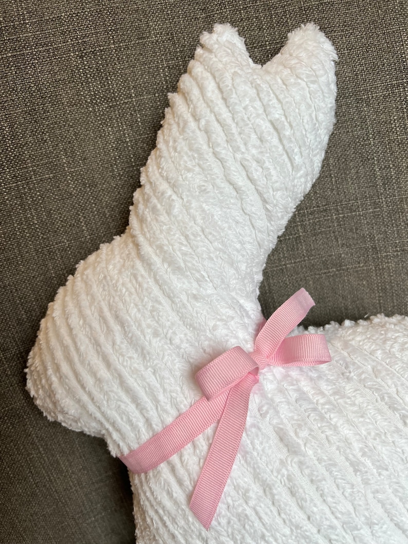 K&ouml;nnte beinhalten: Ein wei&szlig;er Pl&uuml;schhase mit einem rosa Band um den Hals. Der Hase ist aus einem weichen, strukturierten Stoff.