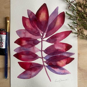 May include: A watercolour painting on white paper featuring a central botanical leaf structure with overlapping petals in shades of deep magenta, crimson, and violet. The artwork includes a signature in the bottom right corner and a paint tube.
