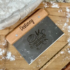 May include: A wooden dough scraper with a stainless steel blade. The blade has a logo that says "Sourdough Mama" and a design of a loaf of bread. The handle is engraved with the name "Destiny".