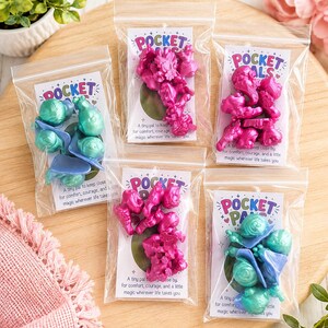 May include: Five clear plastic bags containing small, metallic-finished figurines. Two bags hold teal and blue figures, while three bags hold bright pink figures. Each bag includes a white card labeled POCKET PALS with text about comfort and magic.