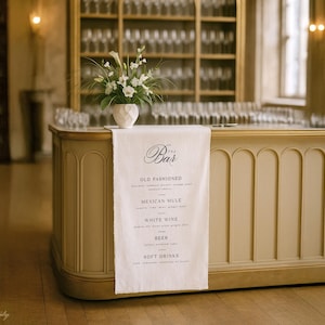 May include: A cream-coloured bar with a white linen runner displaying a menu of drinks. A white vase with white flowers sits on the bar. The bar has a curved front and is in a room with a wooden floor. The menu lists cocktails.