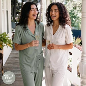May include: Two women wearing matching pajama sets, one in sage green and the other in cream. Both sets include short-sleeved button-down shirts and drawstring pants. Each woman holds a champagne flute. The image is taken outdoors on a wooden porch.