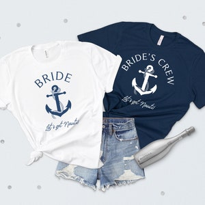 May include: Two white t-shirts with a blue anchor and the text "Bride" and "Bride's Crew" with the phrase "Let's get Nauti!" on each. The shirts are laid out on a white surface with a pair of denim shorts and a silver bottle of sparkling wine.