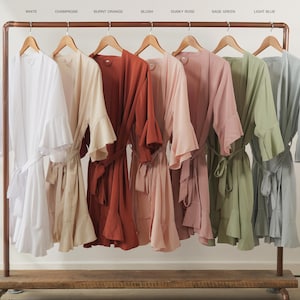 May include: A clothing rack displays six robes in various colors: white, champagne, burnt orange, blush, dusky rose, sage green, and light blue. Each robe has wide sleeves and a matching belt.