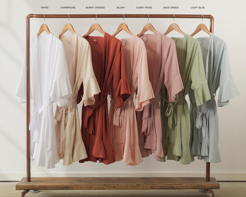 May include: A clothing rack displays six robes in various colors: white, champagne, burnt orange, blush, dusky rose, sage green, and light blue. Each robe has wide sleeves and a matching belt.